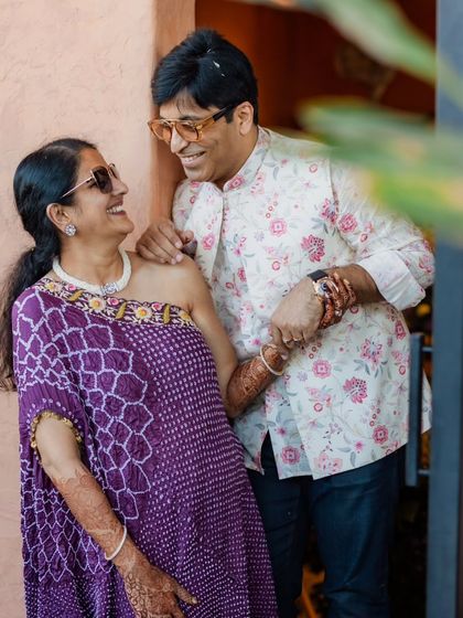 A sweet moment between the parents. Her one-shoulder bandhani top and his floral jacket create a stylish, coordinated couple look that is perfect for a fun, daytime wedding event.
