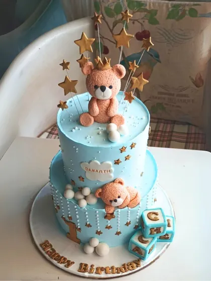 A charming two-tier teddy bear cake in blue, decorated with stars, building blocks, and a cute crowned bear for a first birthday.