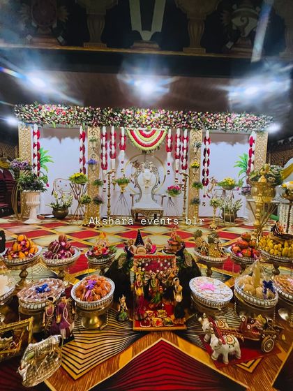 A beautifully lit Seemantham stage at night. The decor features a white backdrop with red and yellow floral hangings, an ornate chair, and a lavish display of offerings on the floor.