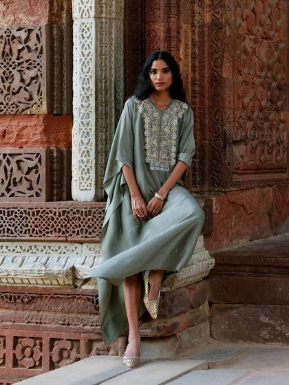 A model sits gracefully in a sage green kaftan with an embroidered yoke, set against a historic, carved stone backdrop.