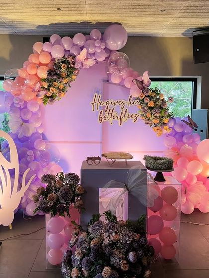 A beautiful butterfly-themed stage decoration. It features a balloon arch in pastel colors with floral accents, a custom "He gives her butterflies" sign, and a large butterfly wing prop.