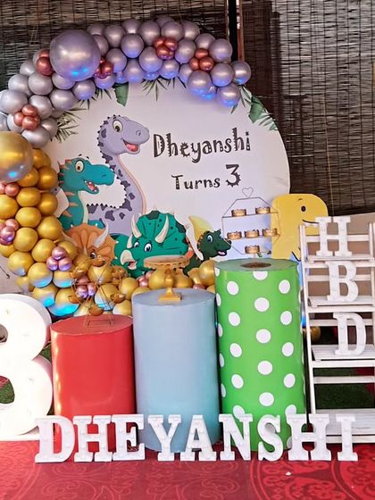 A roaring Dinosaur-themed third birthday party. The setup includes cute dinosaur cutouts, a large light-up number '3', and a 'DHEYANSHI' marquee.