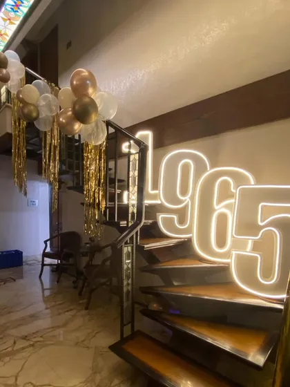 A grand entrance for a 60th birthday, with large LED numbers spelling out the birth year "1965" on a spiral staircase. This dramatic lighting feature, paired with gold-themed balloons, makes for a stunning first impression.