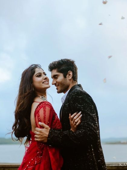 A candid moment of connection as petals fall around the couple during their lakeside pre-wedding session. The woman's vibrant red gown stands out beautifully against the soft sky.