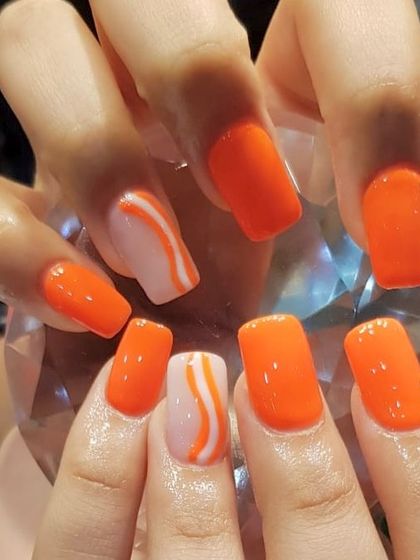 Manifesting summer with these bright orange nails. This set features a bold, glossy orange polish with accent nails showcasing a cool white swirl design.