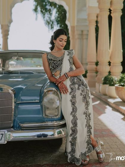 A stylish portrait of the bride leaning against a vintage blue car. Her elegant white and black sari and confident pose create a chic and timeless image.