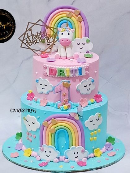 A delightful two-tier unicorn and rainbow cake. The pink and blue tiers are adorned with smiling clouds, colorful rainbows, and a cute unicorn topper for a happy first birthday.