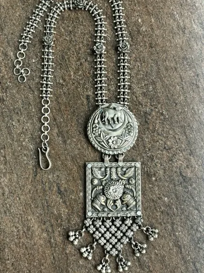 A simple yet stunning oxidised silver necklace. This piece features two pendants, one with an elephant motif and one with a square design, both with intricate details and dangling silver beads.