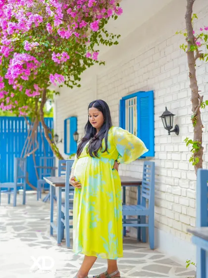 A beautiful solo shot of the mother-to-be in a flowing yellow dress, set against a white brick wall with blooming flowers.