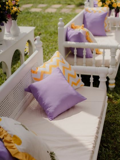 The seating arrangement for guests, featuring white wooden benches with custom cushions in purple, yellow chevron, and sunflower prints, perfectly matching the Haldi theme.