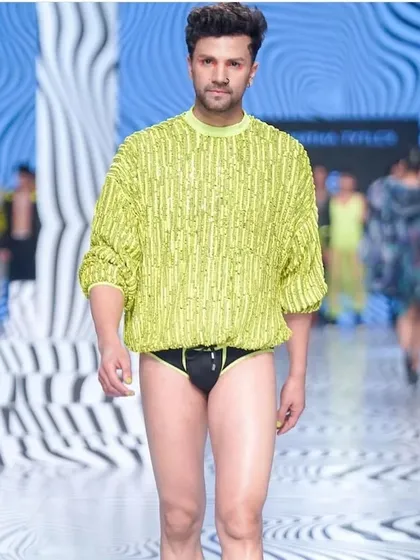 A men's runway look from LFW featuring a clean complexion and subtle grooming. The makeup ensures the model looks fresh and polished under the bright lights, complementing the avant-garde fashion.