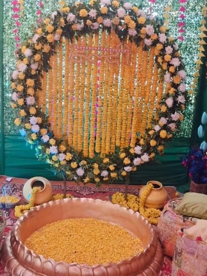 A cozy Haldi setup with a large brass urli and low gadda seating with printed cushions. The backdrop is a beautiful ring of marigolds and white flowers against a green drape.