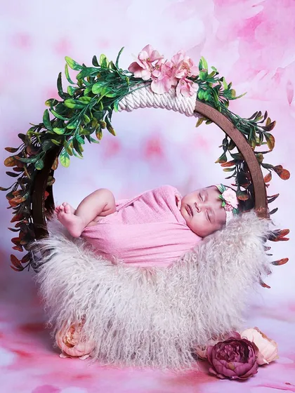 A baby girl wrapped in pink, sleeping on a soft fur cushion inside a round floral hoop prop. The pink floral background creates a dreamy, cohesive look.