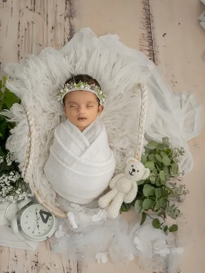 An angelic overhead shot of a newborn baby, swaddled in pure white fabric and resting in a basket adorned with fresh greenery and a tiny teddy bear.