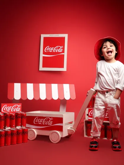 A young boy shows off his personality in a fun, Coca-Cola themed photoshoot. Dressed in a red hat and white outfit, he laughs while posing with a branded cart and cans, demonstrating a creative and vibrant approach to kids' photography.