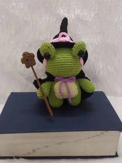 The little wizard frog amigurumi sitting on his book of spells. The details like the purple ribbon and flower on his wand make him extra special.