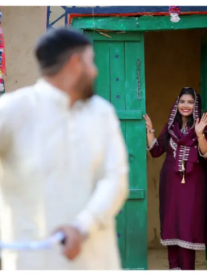 A playful shot from the village-themed pre-wedding shoot. The groom is out of focus in the foreground, creating a sense of depth and drawing attention to the bride's happy wave.