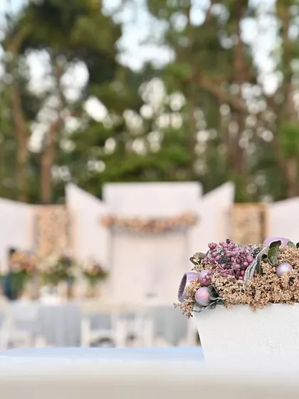 A simple yet elegant centerpiece with dried lavender and pastel flowers on a guest table, with the outdoor wedding stage blurred in the background.