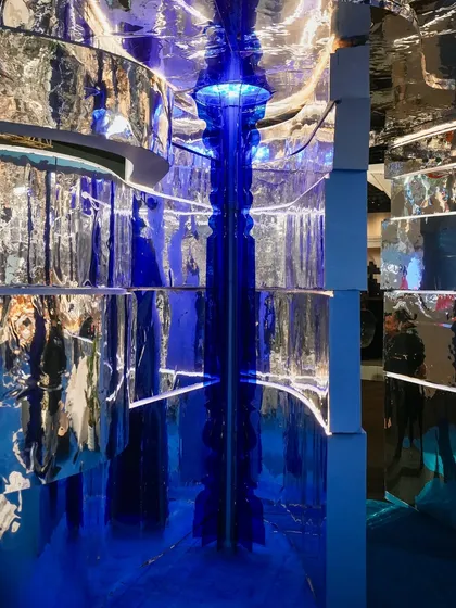 A vertical view looking up through the heart of the installation. The glowing blue column pierces through the space, reflected infinitely in the chrome ceiling, creating an illusion of endless height and depth.
