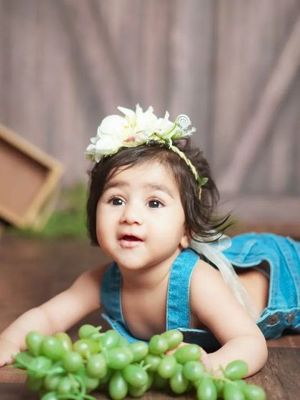 A six-month-old baby girl in a rustic barn wood setting. The contrast of the denim overalls and floral headband is adorable.