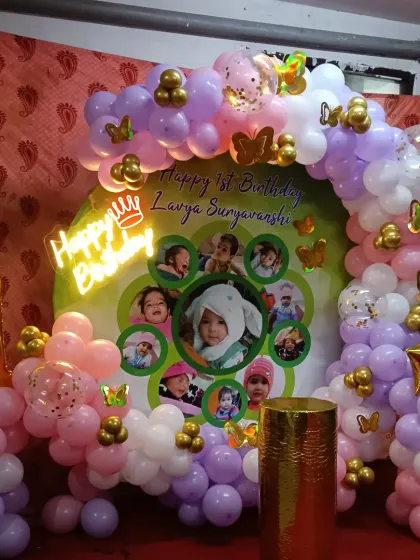 A personalized first birthday with a custom photo collage backdrop, a "1" foil balloon, and a pastel pink and purple balloon arch.