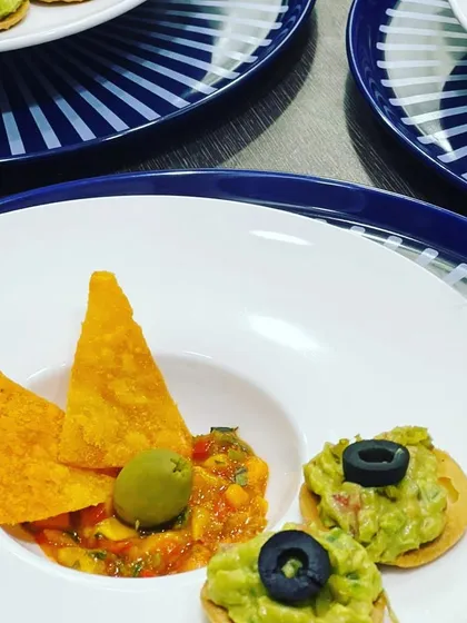 A plated appetizer course featuring a deconstructed nacho bowl with guacamole, salsa, and a crispy tortilla chip.