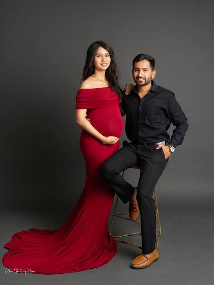 A classic and poised couple's portrait. The expecting mother in a vibrant red gown stands beside her seated partner, creating a timeless image against a simple grey studio background.