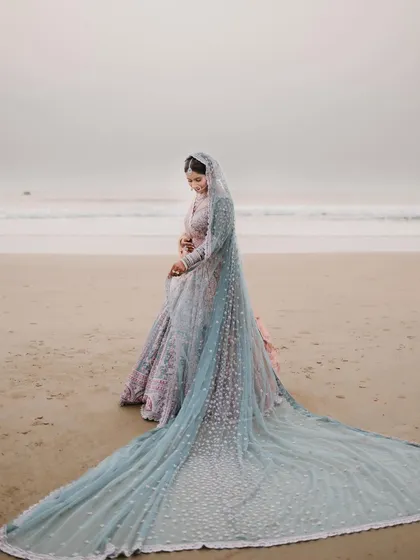 A bride stands on a serene beach at dusk, her magnificent light blue veil trailing behind her, creating a breathtaking and ethereal bridal portrait against the ocean.
