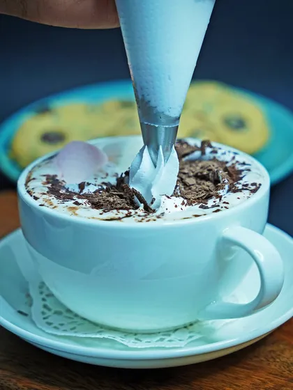 An action shot capturing whipped cream being piped onto a cup of hot chocolate. This dynamic image makes the beverage feel fresh and indulgent, perfect for a cafe's promotional materials.
