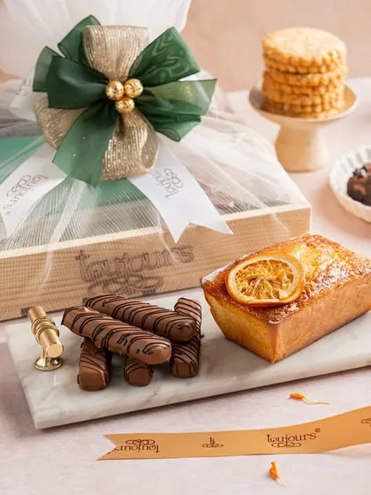 A stylish gift set featuring a lemon tea cake, chocolate-covered sticks, and assorted brownies, all elegantly arranged around a wrapped wooden hamper. This shows a modern and fresh take on Diwali gifting.