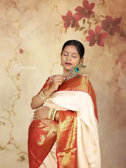 A serene studio portrait of a mother-to-be in a traditional red and white saree, holding a peacock feather.