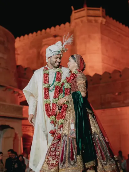 Against the historic walls of Suryagarh fort, the couple shares a look of love after their varmala ceremony. The rich colors of their attire stand out against the warm, illuminated stone, creating a timeless portrait.