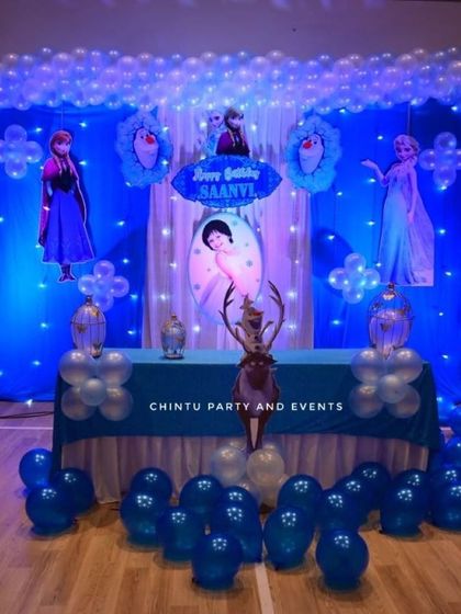 A classic Frozen theme birthday party with a full stage setup. The decor uses blue and white drapes, character cutouts of Elsa and Anna, and a balloon arch to create a winter wonderland.