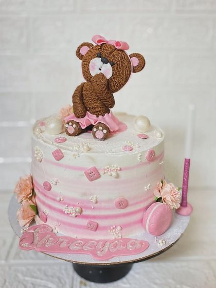 A beautiful pink and white cake with a detailed, hand-crafted teddy bear topper.
