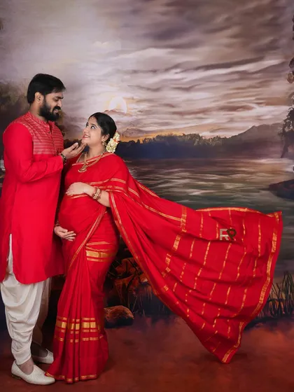 This portrait captures a couple in matching red traditional outfits against a painted landscape backdrop. The flowing saree adds a touch of drama and elegance to the scene.