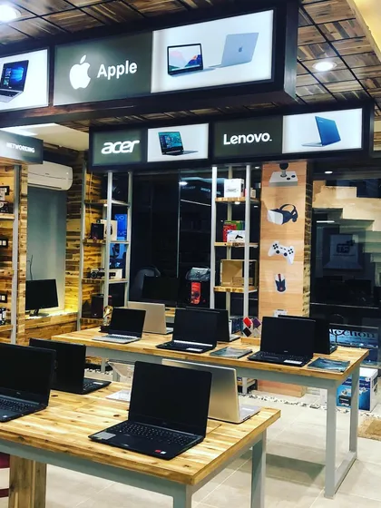 This view of the computer showroom highlights the organized layout, with laptops displayed on large wooden tables for easy access. The walls are lined with shelving for accessories, maximizing vertical space.