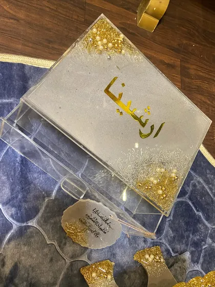 A personalized gift set featuring a dome-shaped prayer mat in a beautiful blue pattern. The set is completed with a custom resin Quran box and holder, a crystal tasbeeh, a bookmark, and a car hanging, all in a coordinated white and gold glitter design.