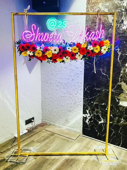 A creative photo frame for a party, featuring a neon sign with the couple's names and a colorful arrangement of gerbera daisies.