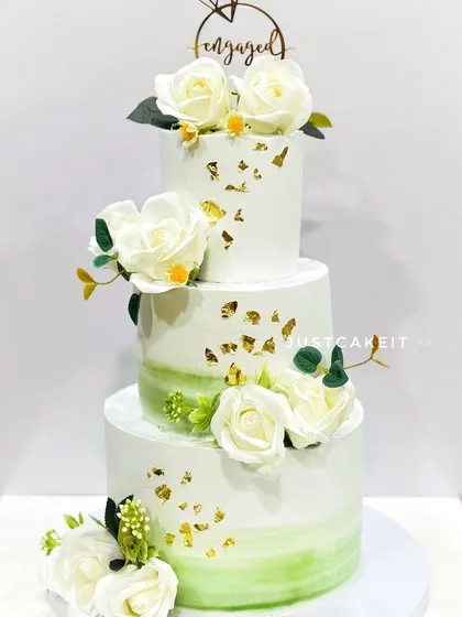 A three-tier engagement cake with a soft green watercolor effect and gold leaf. The arrangement of white sugar roses adds a classic, romantic touch.