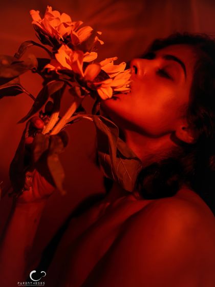 A sensual and artistic pose, breathing in the scent of the sunflower. The red light envelops her, creating a warm and intimate atmosphere.