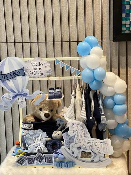 The 'Hello Baby Dhruv' hamper features a mini wardrobe filled with clothes, a teddy bear, and a rocking horse, all decorated with a beautiful blue and white balloon arch.