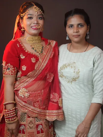 A proud student stands with her model, who is styled as a North Indian bride in a red lehenga, after a successful class practical.