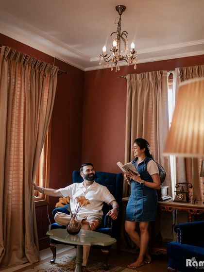 A candid moment captured in a vintage-style room. The groom looks out the window while the bride reads a book, creating a scene that feels natural and tells a story about their shared life.