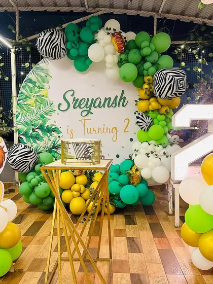 A jungle themed second birthday party on a terrace. The decor includes a backdrop with tropical leaves, a lion cutout, zebra-print balloon accents, and a large light-up number '2'.