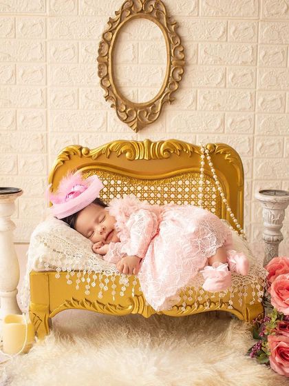 A royal nap. This newborn sleeps on an ornate golden chaise lounge, dressed in a pink lace dress and a tiny hat.