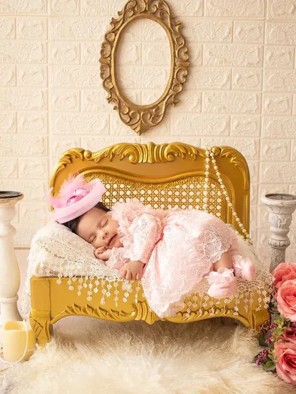A royal nap. This newborn sleeps on an ornate golden chaise lounge, dressed in a pink lace dress and a tiny hat.