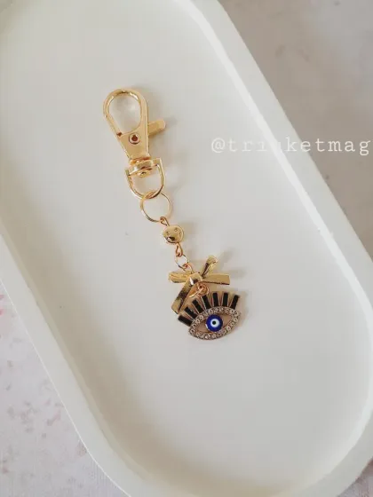 A chic and protective evil eye charm with a bow, perfect for attaching to your bag, keys, or lip balm. The gold and crystal details add a touch of glam.