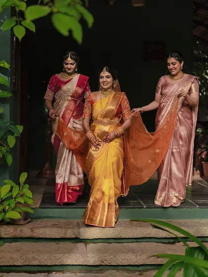 The bride, in a beautiful golden-yellow saree, is helped by her sisters as she walks.