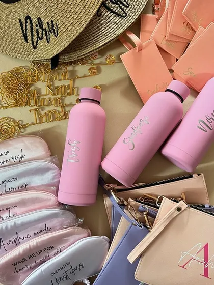 A collection of goodies ready to be packed into a beach bachelorette hamper. Featuring personalised water bottles, eye masks, pouches, and a custom sun hat.