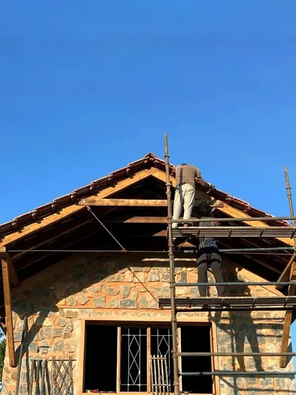 Building is a human endeavor. Here, craftsmen work under the open sky, laying tiles on a roof supported by a robust wooden frame and solid stone walls. This image captures the strength of natural materials and the dedication that goes into every home we build.